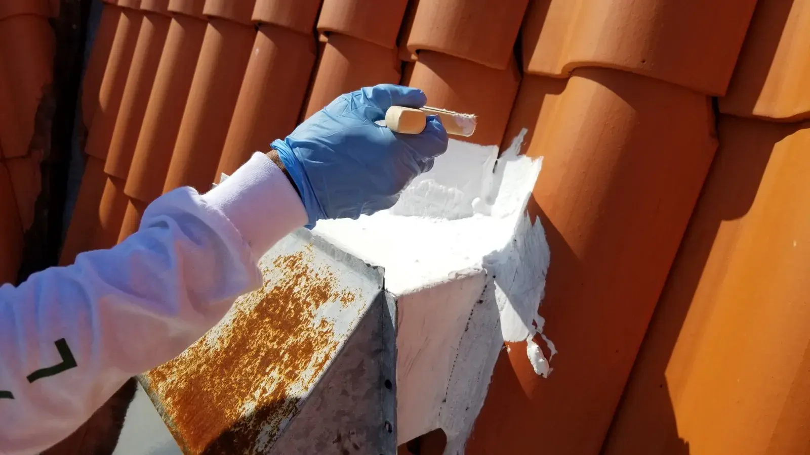 A person in gloves applies white paint to a roof's corner, showcasing vibrant orange tiles and careful detail work. 